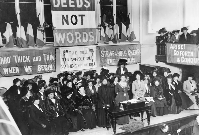 Die Abbildung zeigt ein Treffen von Frauen der Organisation „Women’s Social and Political Union“ in Manchester im Jahr 1908. Zu sehen ist ein großes Plakat mit den Worten „Deeds not Words“, übersetzt „Taten statt Worte“.